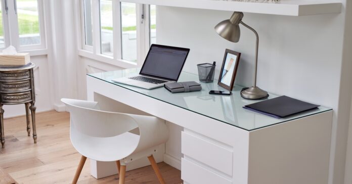 An organized white desk features an open laptop, a silver lamp, a picture frame, a pen holder, a smartphone, and a file.