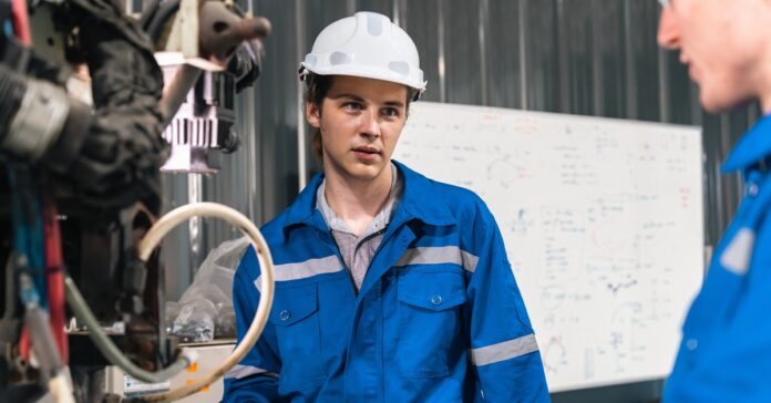 Two engineers wearing blue outfits looking at a large machine in a brightly lit industrial workplace.