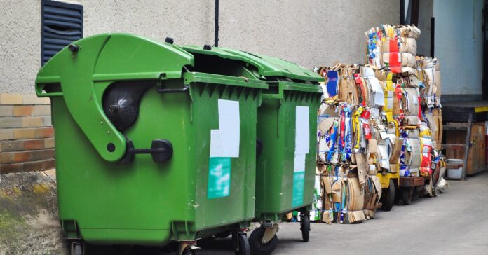 An exterior wall at a business with two large green trash bins next to stacks of bundled cardboard.
