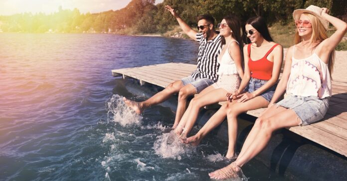Four adults sitting on a wooden dock with their feet in the water beside a tree-lined body of water.
