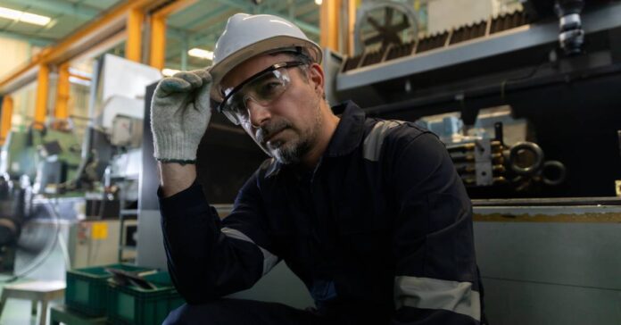 A man wearing a white hard hat, safety glasses, and work gloves is sitting down near large industrial machines.