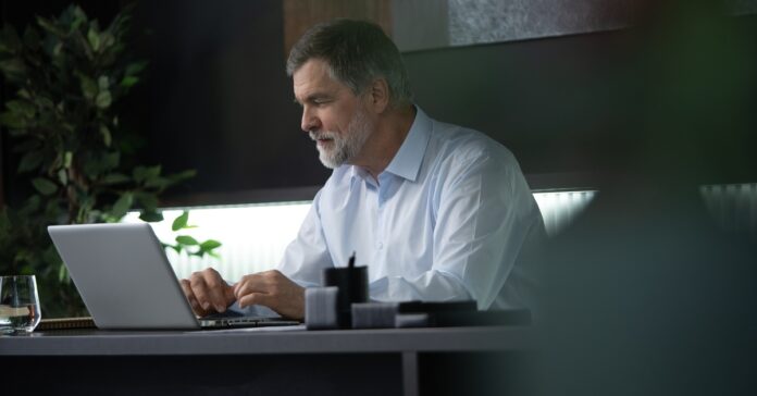 A businessman sits in his office at work and looks at his laptop. He smiles and wears a blue, button-down shirt.