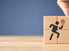 A hand placing a wooden block onto a four-block stack that has a picture of a running figure holding a light bulb.