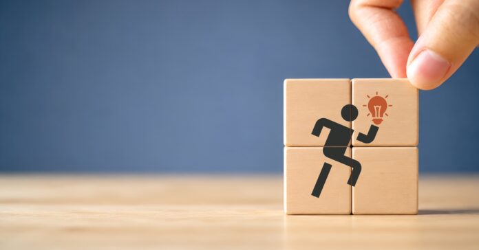 A hand placing a wooden block onto a four-block stack that has a picture of a running figure holding a light bulb.