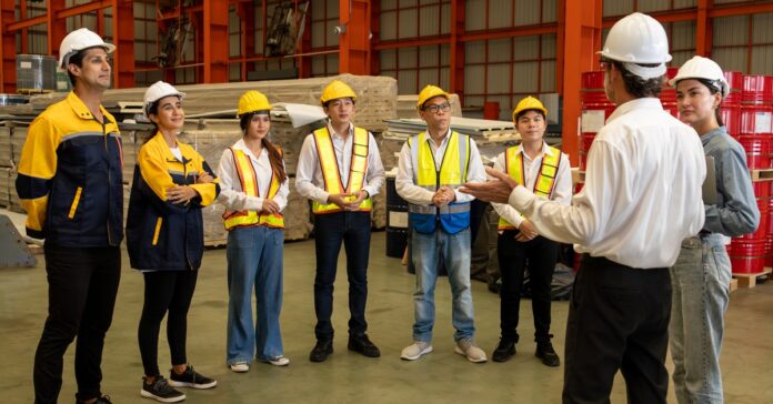 A manager stands in front of his employees in a warehouse and talks to them. The workers all wear hard hats.