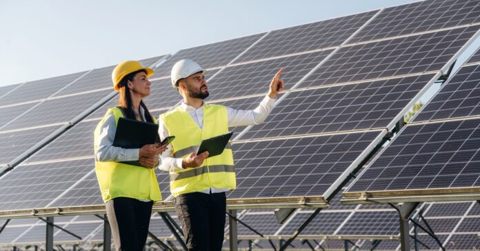 Two solar engineers reviewing a large-scale solar project. Both engineers are wearing safety gear and holding tablets.