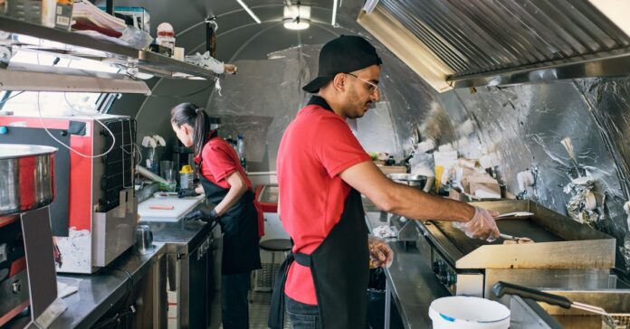 Two workers stand on opposite sides of a food truck. One worker fries meat while the other preps and orders.