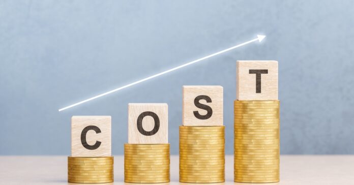 Wooden blocks stand on stacks of coins, spelling out the word Cost. A white arrow floats in the air above them.