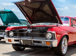 a red classic car and a turquoise classic car parked outdoors with open hoods under a partly cloudy sky.
