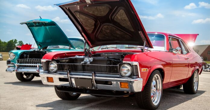 a red classic car and a turquoise classic car parked outdoors with open hoods under a partly cloudy sky.