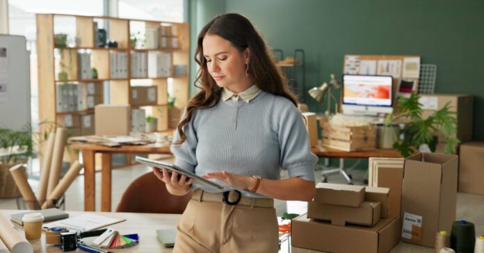 A businesswoman using a tablet stands beside cardboard boxes in a bright office with desks, shelves, plants, and supplies.