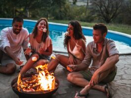 How To Plan a Backyard Around Swimming and Entertaining Four friends roasting marshmallows around a fire pit while sitting next to an outdoor swimming pool during sunset.