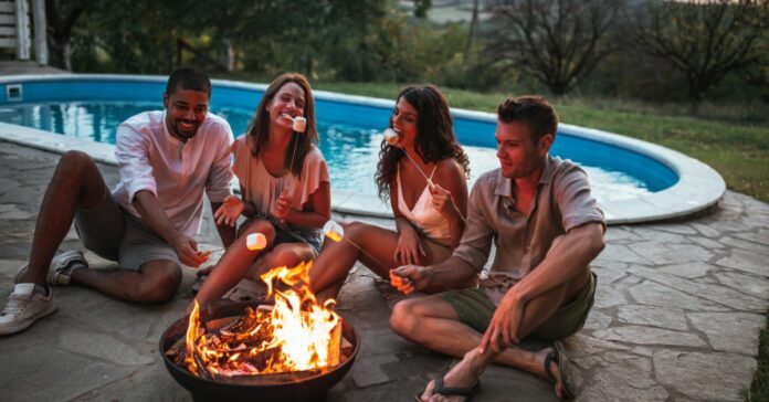 Four friends roasting marshmallows around a fire pit while sitting next to an outdoor swimming pool during sunset.