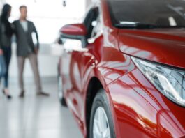 A blurry couple standing behind a red car in a showroom, looking at it. The vehicle is shiny and brand new.