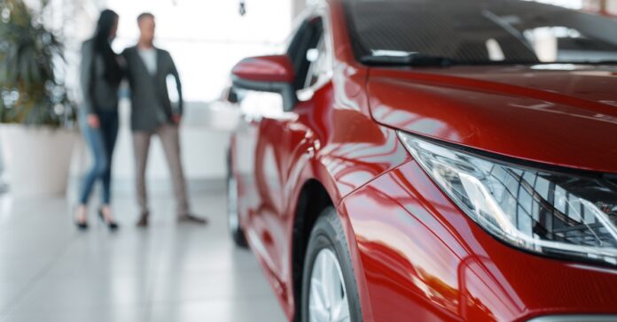 A blurry couple standing behind a red car in a showroom, looking at it. The vehicle is shiny and brand new.