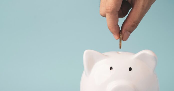 A close-up view shows a hand pinching a coin between its fingers and holding it above the opening of a white piggy bank.
