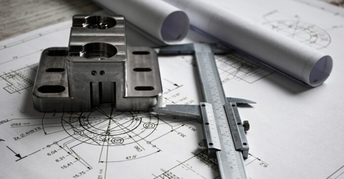 A metal machined component and a vernier caliper resting on engineering drawings beside rolled blueprints.
