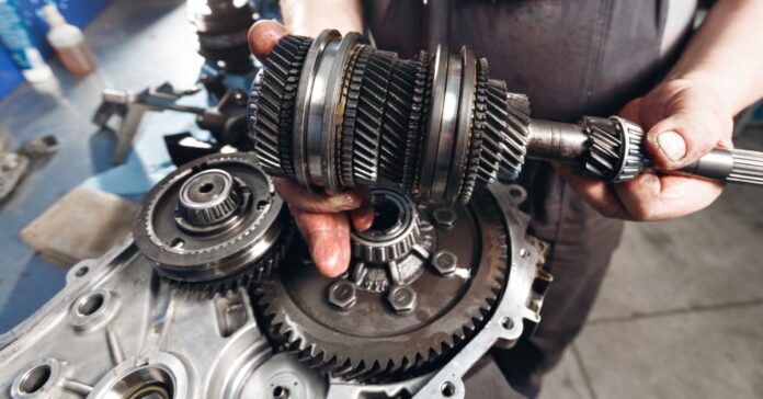 A close-up view shows a man holding gears from inside a gearbox. Other metal gears sit on the table underneath.