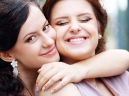 Person wearing floral earrings embraces another person in a mauve dress, both smiling at an outdoor celebration.