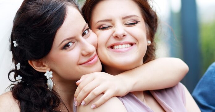 Person wearing floral earrings embraces another person in a mauve dress, both smiling at an outdoor celebration.