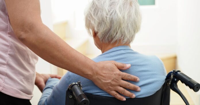 The back of a person's head with grey hair as they sit in a wheelchair. Someone else has their hand on their back.