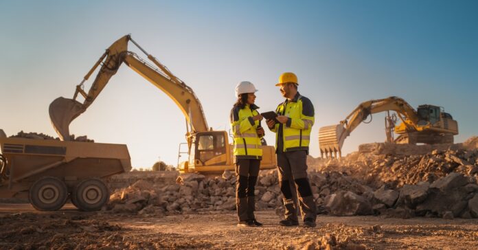 Two people wearing high-visibility jackets and hard hats are standing and talking on a jobsite with excavators.