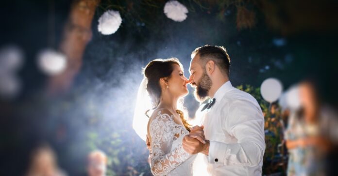 A newlywed couple dancing closely outdoors at night, lit from behind, with blurred guests and round lights nearby.