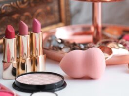 Close-up of three golden lipsticks, a blending sponge, a contouring palette, and some jewelry on a white surface.