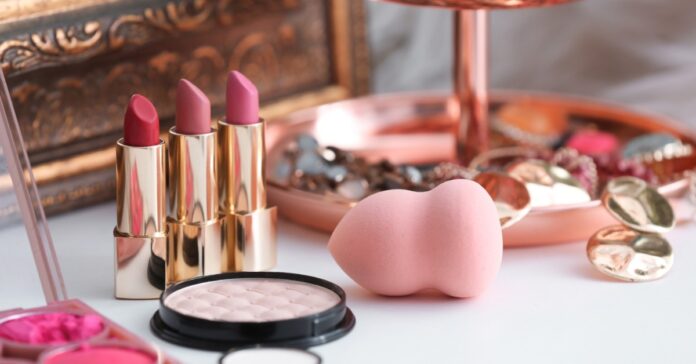 Close-up of three golden lipsticks, a blending sponge, a contouring palette, and some jewelry on a white surface.