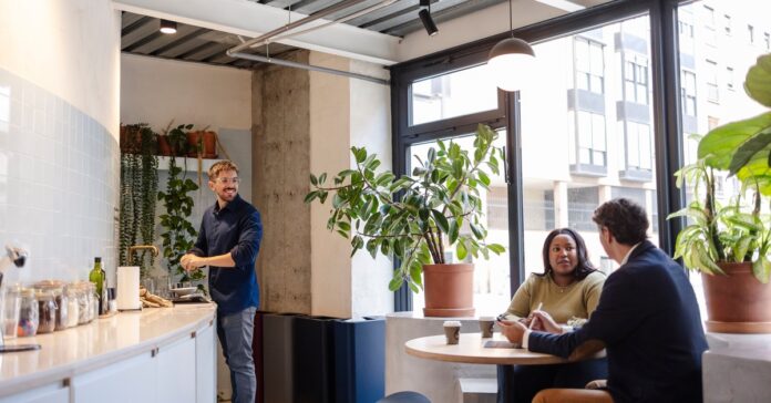 Three coworkers gathered in a bright office break room, with two seated at a table and one standing by the sink.