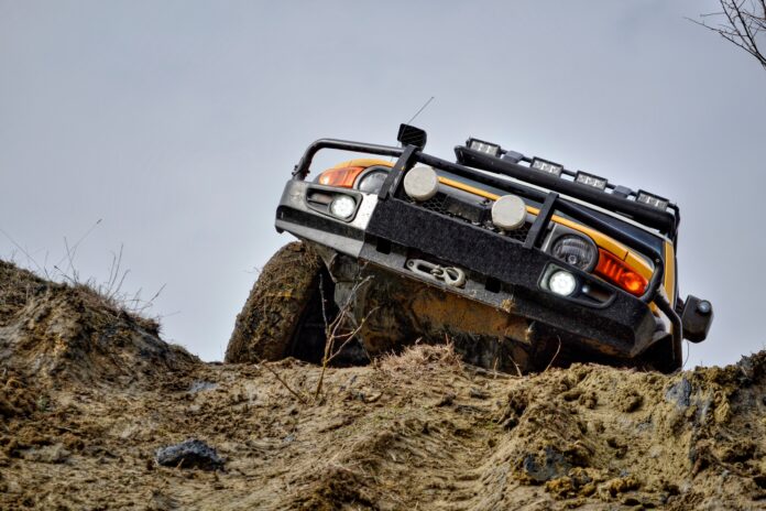 A yellow Bronco with metal protection and bar lights on top, climbing a small hill through a dirt road covered in mud.