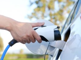 How To Reduce Fire Risks From EV Batteries A person is holding a charging cord near the charging port on a white electric vehicle that's outside.