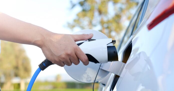 A person is holding a charging cord near the charging port on a white electric vehicle that's outside.
