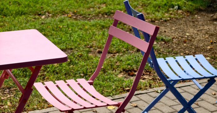 Red and blue metal patio chairs sit beside a matching table on a paved outdoor area surrounded by green grass.