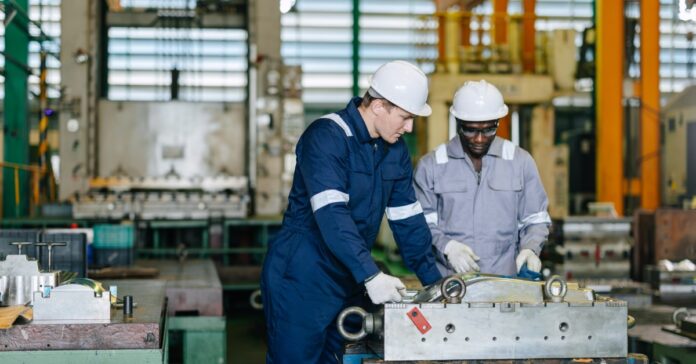 Two engineers wearing white hard hats and white gloves look at a metal machine in a large manufacturing plant.