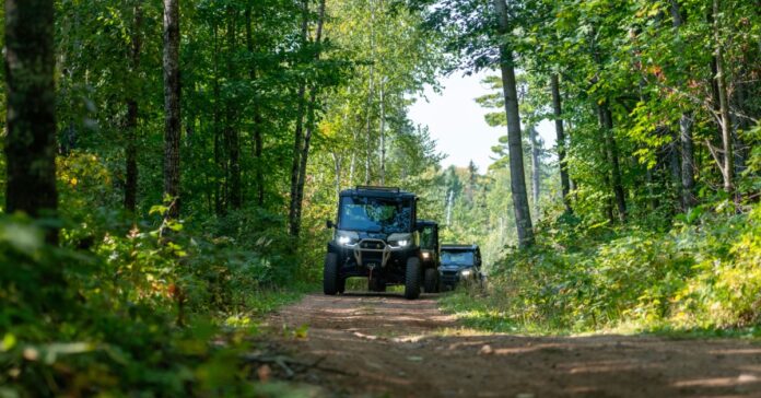 An all-terrain vehicle driving along a dirt trail through a wooded area with tall trees and green foliage.
