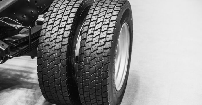A close-up of the frame of a dually pickup truck with two black tires parked on a white floor with a white background.