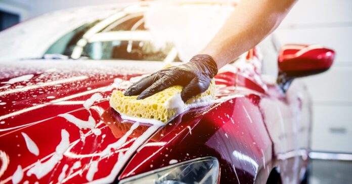 A person wearing black gloves while washing the hood of a red car with a yellow sponge covered in white soap suds.