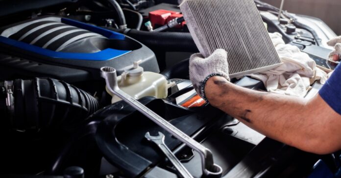 A person wearing work gloves holding a dirty rectangular air filter above an open car engine with tools nearby.