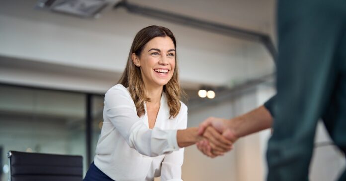 A smiling woman wearing professional business attire reaches across the table to shake someone's hand.