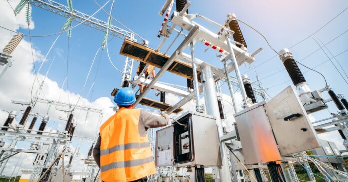A technician wearing a high-visibility vest and hard hat working on a small panel in an outdoor electrical station.