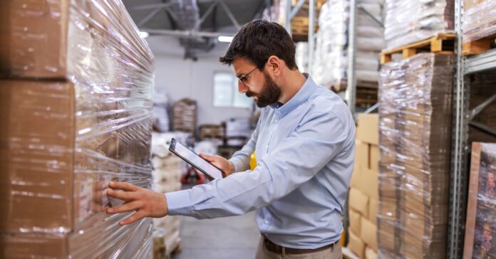 A warehouse supervisor uses a tablet while examining shrink-wrapped boxes stacked on pallets in a storage facility.