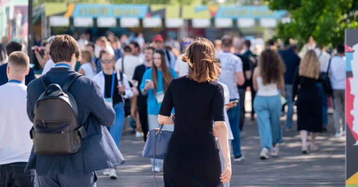 A woman in a black dress carrying a handbag walks among a crowd of people along a tree-lined outdoor path.