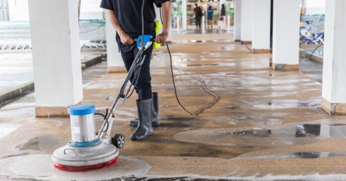 A worker operates a commercial floor-cleaning machine on a wet concrete walkway beneath a covered building entrance.