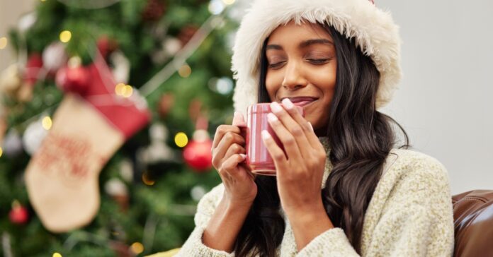 A young woman sits on the couch cozy with a warm coffee mug in both hands. She wears a Santa hat with a tree behind her.