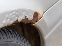 Risks of Rust in the Winter: How To Care for Your Vehicle The white body panel of a car with bubbling paint and a jagged patch of reddish-brown rust above a black tire and wheel arch.