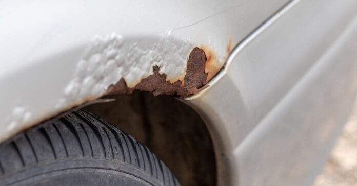 The white body panel of a car with bubbling paint and a jagged patch of reddish-brown rust above a black tire and wheel arch.
