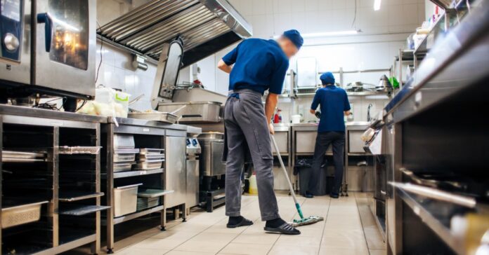 An employee mopping the tile floor in a commercial kitchen. Another employee works at a sink nearby.
